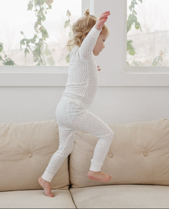 Child in tealbee pajamas in dash print standing on a beige sofa near a window with natural light.