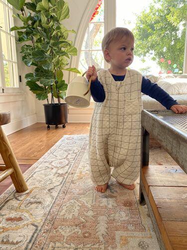 Child in a checkered tealbee dreamsuit holding a mug in a home setting with plants and furniture.