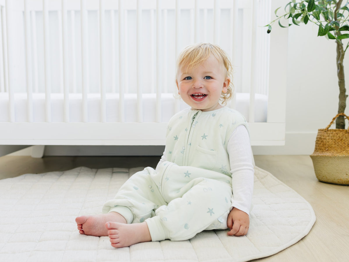 Toddler in a Seashell Dreamsuit sitting on a rug by a crib, laughing